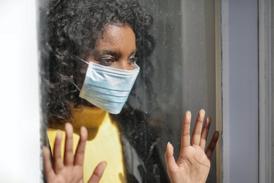 A female wearing a facemask and standing with her hands placed against a window. She has an unhappy expression. A female wearing a facemask and standing with her hands placed against a window. She has an unhappy expression.