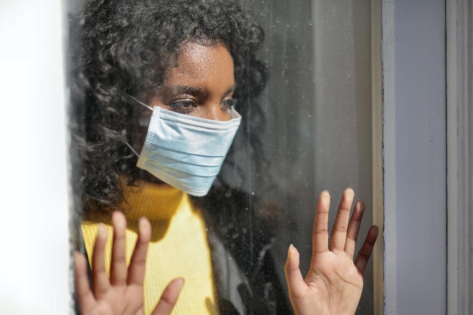 A female wearing a facemask and standing with her hands placed against a window. She has an unhappy expression. 